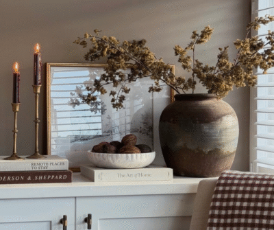 Table styled with natural elements including green foliage, books, faux wax candles, and a black-and-white print, creating a balanced and inviting home decor vignette.